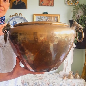 May include: A large, polished brass bowl with a wide, rounded body and a smooth, reflective surface. The bowl features two decorative metal handles on opposite sides, each shaped like an animal head with a circular ring hanging from its mouth.