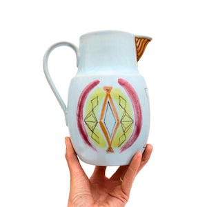 May include: A light blue ceramic pitcher with a curved handle and a spout with brown stripes. The pitcher features a colorful geometric design with red, yellow, and orange accents. The pitcher is held by a hand.