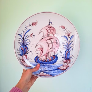 May include: A decorative ceramic plate featuring a sailing ship with pink sails and a blue hull, set against a blue and white ocean scene. The plate is adorned with blue and red floral accents and a red rim.