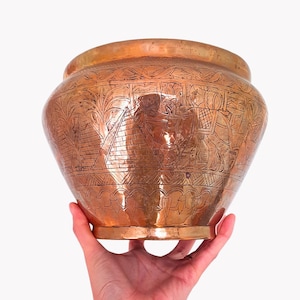 Könnte beinhalten: Eine kupferfarbene, runde Vase mit aufwendigen Ätzmustern. Die Vase hat eine breite Öffnung und ein detailliertes Muster, das Pyramiden, Figuren und andere ägyptische Motive darstellt. Die Vase wird von einer Hand gehalten.