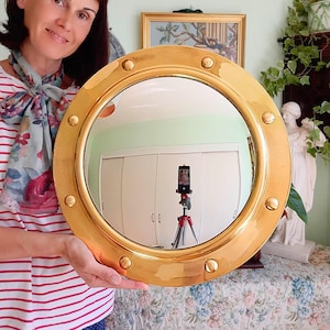 May include: A circular porthole-style mirror with a polished gold-tone metal frame. The frame features six evenly spaced decorative rivets around the perimeter. The reflective glass surface is clear and circular, set within the thick, smooth metal border.