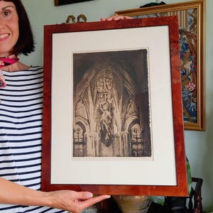 May include: A framed art print featuring a dark, monochromatic etching of a gothic architectural interior with vaulted ceilings and intricate stone carvings, encased in a polished, reddish-brown burl wood frame with a wide cream-colored mat.