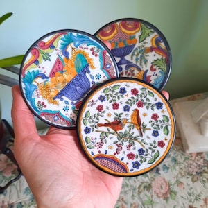 May include: Three round ceramic coasters with colorful floral and fruit designs. One coaster features two birds on a branch. The coasters have black rims and are held in a hand.