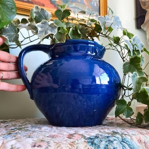 May include: A deep cobalt blue ceramic pitcher with a rounded, bulbous body and a single curved handle. The pitcher features a smooth, glossy finish and a wide opening at the top, providing a solid vessel for holding liquids or decorative items.