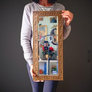 May include: A rectangular gold-framed mirror with decorative floral and leaf patterns. The mirror reflects a room with artwork, a shelf with books and flowers, and a ring light with a phone attached. The frame is held by a person.