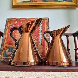 May include: Two copper-coloured jugs with black handles are displayed on a wooden surface. The jugs have a sleek, modern design with a pointed spout. The background includes a decorative rug and a framed artwork, adding to the vintage aesthetic.