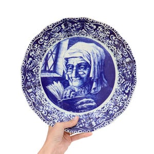 May include: A blue and white decorative plate with a detailed illustration of an elderly woman reading. The plate has a scalloped edge with a floral pattern. The woman wears a head covering and spectacles.