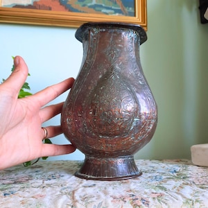 Könnte beinhalten: Eine Kupfervase mit bauchigem Körper und ausgestelltem Rand. Die Oberfläche ist mit komplexen, geprägten Blumen- und Geometriemustern verziert und weist eine verwitterte, oxidierte Patina mit grünen und dunkelbraunen Flecken auf dem Metall auf.