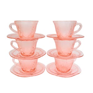May include: Six pink glass teacups and saucers are stacked in pairs. The cups and saucers have a delicate, embossed floral design. The cups have handles and are set on matching round saucers. The set is against a white background.