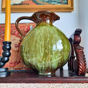 May include: A large, bulbous ceramic jug with a brown top and handle, transitioning to a green body with vertical streaks. The jug sits on a dark wooden surface, reflecting the light. A decorative piece.