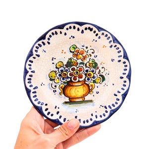 May include: A hand-painted ceramic plate with a floral design. The plate features a cream-colored background with a blue scalloped edge. A gold vase filled with colorful flowers is in the center.
