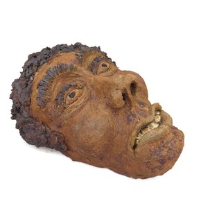 May include: A ceramic sculpture of a human face with a distressed brown finish. The face is contorted in a grimace, with the mouth open and teeth showing.
