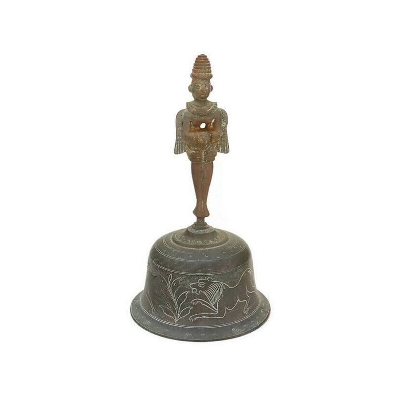 Op de afbeelding: Een bronzen bel met een decoratief handvat met een staand figuur. De bel heeft een ronde basis met een gegraveerd ontwerp van een leeuw en bladeren.
