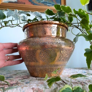 May include: A polished copper pot featuring intricate hand-engraved patterns, including geometric borders and circular motifs with detailed scrollwork. The vessel has a wide, rounded body that tapers towards the top rim, showcasing a warm metallic finish.