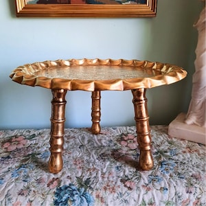 May include: A round gold-toned metal pedestal tray featuring a scalloped, ruffled edge and a flat surface with an intricate engraved pattern. The tray is supported by three sturdy, segmented legs that resemble bamboo stalks in a polished finish.