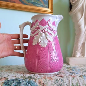 May include: A ceramic jug featuring a vibrant pink body with a textured wood grain pattern. The exterior is decorated with white relief accents depicting oak leaves and acorns arranged in a cross pattern. It has a white handle and rim.