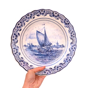 May include: A white ceramic plate with a blue and white design of a sailboat and windmills. The plate has a scalloped edge and a floral pattern.