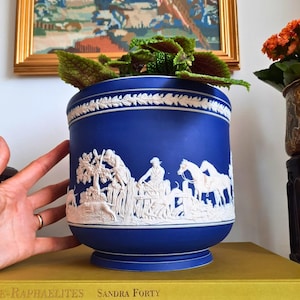 May include: A blue ceramic planter with white relief detailing depicting a hunting scene. The planter has a wide rim and a pedestal base. The planter is filled with a green and red plant. A hand is visible on the left side of the planter.