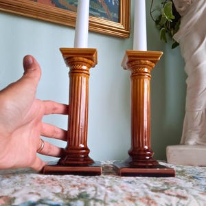 May include: Two ceramic candle holders shaped like classical columns with a glossy brown gradient finish. Each holder features a square base, a fluted shaft, and a square top platform, currently holding a tall, slender white taper candle.