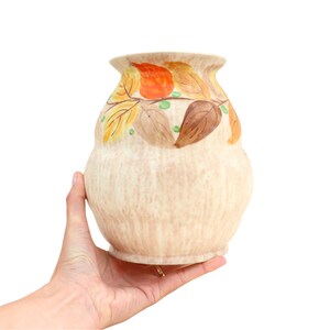 May include: A cream-coloured ceramic vase with a hand-painted design of brown, orange, and yellow leaves. The vase has a textured surface and a rounded shape.