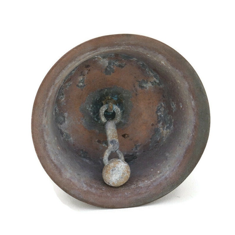 Op de afbeelding: Een vintage, koperkleurige bel met een versleten, verweerde afwerking. De bel heeft een metalen lus en een kleine, ronde knop aan het einde van de lus.