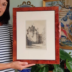 May include: A framed black and white etching of a castle with multiple turrets and a flag on top. The artwork is set within a cream-colored mat and a polished, reddish-brown burl wood frame. The print features detailed architectural lines.