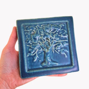May include: A square, ceramic tile with a raised relief of a tree design. The tile is a deep teal colour with a glossy finish. The tree has detailed branches and leaves. The tile is held in a hand.