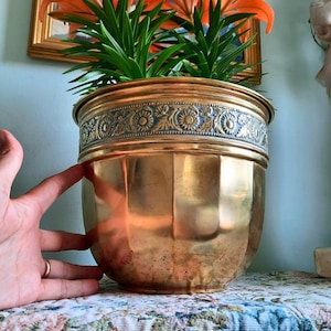 May include: A polished brass planter featuring a wide, embossed band around the upper rim with a repeating floral and leaf pattern. The body of the planter has a smooth, vertical fluted design with a warm, metallic finish and a rounded base.