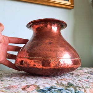 May include: A wide-bodied copper vessel with a rounded shape and a flared rim. The surface features a hammered texture with visible signs of age and a horizontal seam running around the center of the body, showcasing a warm metallic finish.
