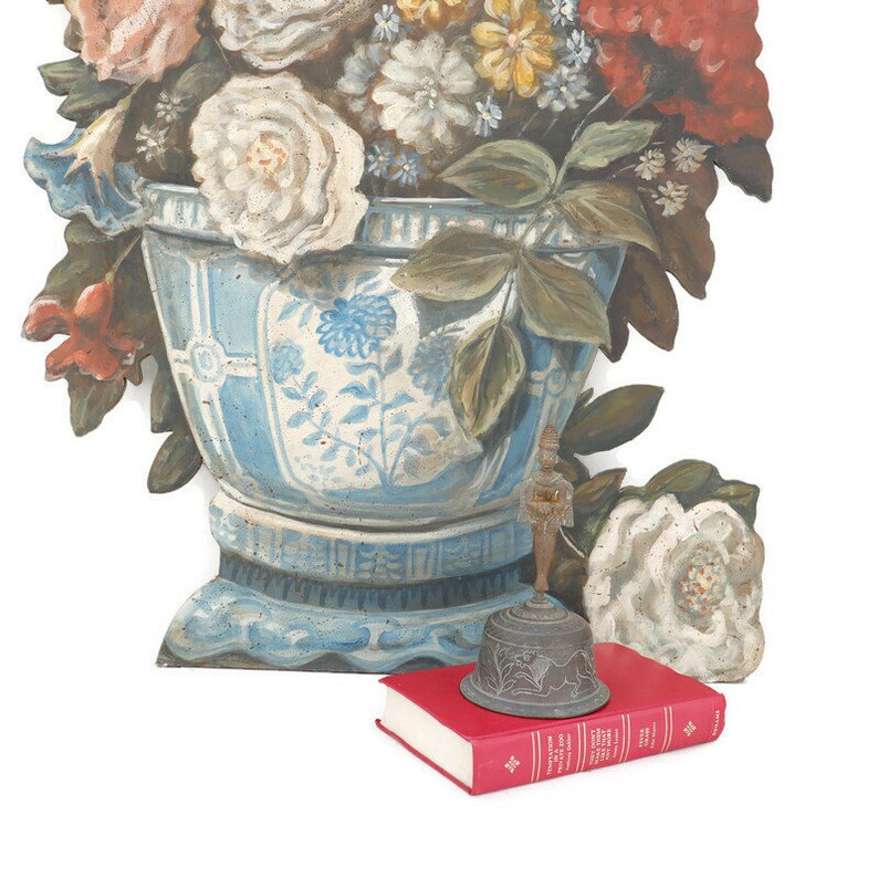 Op de afbeelding: Een blauwe en witte keramische vaas met een bloemenmotief, gevuld met geschilderde bloemen. Een kleine, sierlijke, bronzen bel zit op een rood boek.