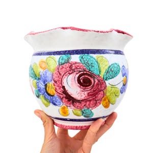 May include: A white ceramic plant pot with a ruffled edge and hand-painted floral design. The pot features a large red rose, blue and yellow flowers, and green leaves. A blue stripe encircles the pot, and the rim is trimmed in red.