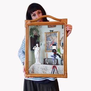 May include: A rectangular, light brown rattan mirror with a curved top handle. The mirror reflects a room with artwork, a statue, and a smartphone on a tripod. The frame has a woven texture, and the overall style is vintage.