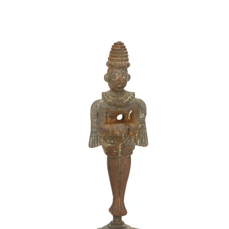 Op de afbeelding: Een klein, bruin, metalen beeldje van een staande figuur met een kegelvormige kroon en vleugels. De figuur houdt zijn handen voor zijn borst bij elkaar.