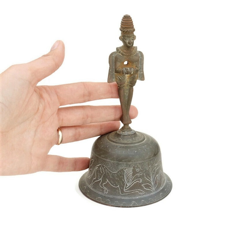 Op de afbeelding: Een kleine, antieke bronzen bel met een gedetailleerd ontwerp. De bel heeft bovenop een figuur van een staande persoon en onderaan een decoratief ontwerp met leeuwen.
