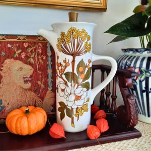 May include: A white ceramic coffee pot with a brown wooden lid and a large handle. The pot is decorated with a floral design in shades of brown, yellow, and green. An orange pumpkin and red seed pods are in the foreground.