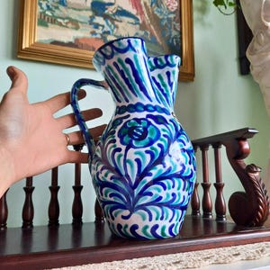 May include: A ceramic jug with a blue, teal, and white floral pattern. The jug has a handle and two spouts. The design features stylized flowers and leaves. The jug is on a dark wooden shelf.