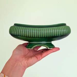 May include: A green ceramic planter with a ribbed design and a pedestal base. The planter has a curved shape and is held in a hand. The planter is a vibrant shade of green.