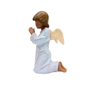 May include: A ceramic figurine of a kneeling angel with brown hair and blue wings. The angel is wearing a light blue robe and has its hands clasped in prayer.
