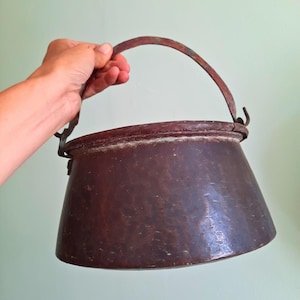 May include: A dark brown, hammered metal pot with a wide, tapered body and a thick, curved handle attached to the sides with metal loops. The surface shows a weathered, textured finish with visible signs of age and oxidation throughout the metal.
