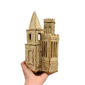 May include: A hand-crafted ceramic sculpture of a building with a pointed roof and a tower. The building is made of white clay and has brown accents. The sculpture is detailed with windows and a door.