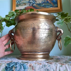 May include: A round brass planter featuring intricate etched patterns of clouds and stylised creatures. The vessel has a wide rim, a flared base, and two side handles shaped like animal heads, each holding a circular brass ring for easy handling.