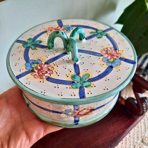 Könnte beinhalten: Ein runder Keramikbehälter mit Deckel, verziert mit einem floralen Muster in Rosa, Blau und Grün. Der Deckel hat einen türkisfarbenen Griff und ist mit blauen Bändern und Blumen verziert. Der Behälter wird in einer Hand gehalten.