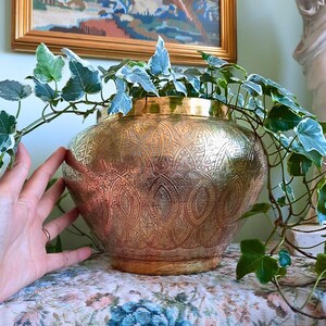 May include: A round, polished brass planter featuring intricate, embossed geometric and floral patterns across its surface. The vessel has a wide, curved body and a smooth, plain rim, currently holding a trailing variegated ivy plant with green and white leaves.