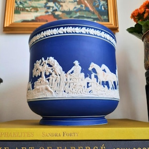 May include: A cobalt blue ceramic vase with a white bas-relief design depicting figures, horses, and dogs in a hunting scene. The vase has a rounded shape and a decorative white border around the top.