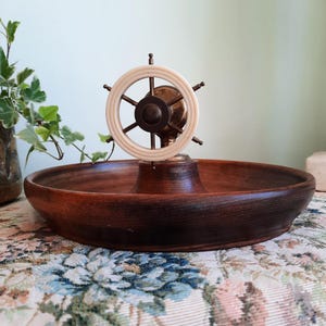 May include: A decorative wooden bowl with a ship's wheel centerpiece. The bowl is dark brown and round, with a raised center supporting the wheel. The wheel is off-white and brown, with a classic nautical design.