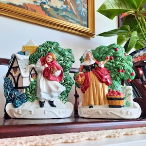 May include: Two ceramic figurines depicting women in traditional attire. One figure wears a red cloak and carries flowers near a snow-covered cottage. The other figure picks apples from a tree, with a basket of apples nearby.