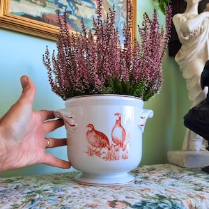 May include: A white ceramic planter with two side handles and a pedestal base, featuring a reddish-orange illustration of two game birds on the front. The planter is filled with a dense cluster of purple heather flowers with green stems.