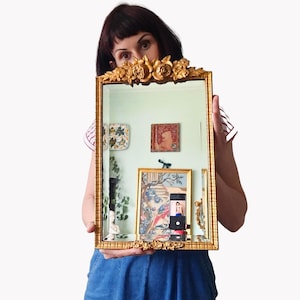May include: A rectangular gold-framed mirror with ornate floral detailing at the top and bottom. The mirror reflects a room with artwork and a smartphone. The frame has a textured, antique appearance.