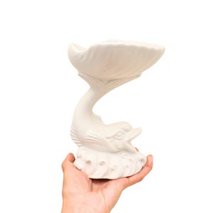 May include: White ceramic dolphin sculpture with a bowl on its back. The dolphin is in a leaping pose with its tail curled up. The bowl is shaped like a seashell.