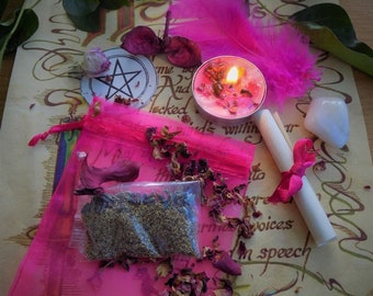 May include: A collection of mystical items, including a pink feather, a lit candle, a pink drawstring bag, a pentagram, and a clear quartz crystal. Dried flowers and herbs are scattered around the items on a parchment-like paper.